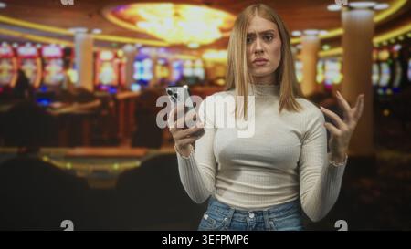 Frau, die ein Smartphone mit einer Hand hält, die Stirn runzelnd, scrollend und die andere Hand in einem hellen Casino-Gebäude hochhebt; frustrierende Kommunikationsprobleme. Stockfoto