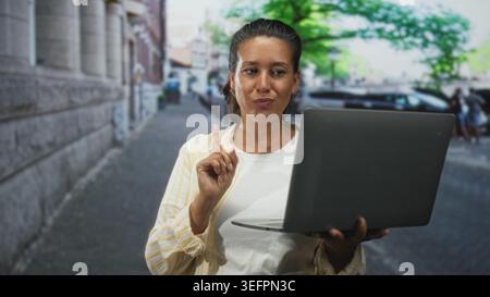 Eine Frau, die den Laptop hält, zeigt den Zeigefinger auf eine urbane Straße, während sie auf den Bildschirm blickt; zielgerichtete Anleitung. Stockfoto