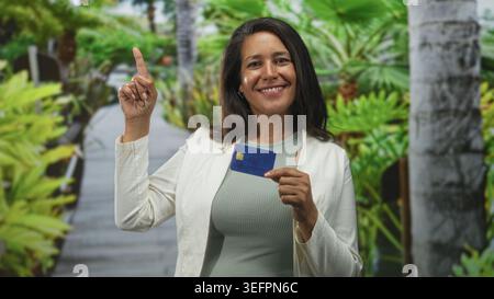Frau, die die Kreditkarte hält, zeigt mit dem Finger nach oben auf der Straße mit tropischen Pflanzen und einer Promenade dahinter; Selbstvertrauen stärken. Stockfoto