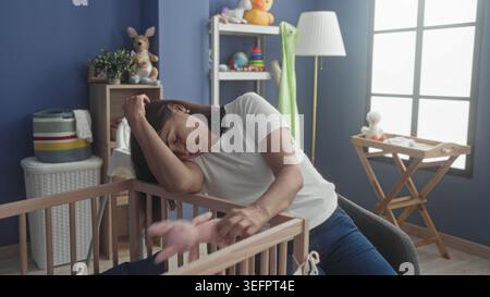 Die Frau legt den Kopf auf das Gittergeländer, übergibt die Augen, hält rosafarbenes Spielzeug im Babyzimmer mit Wickeltisch und Fenster; Pflegemüdigkeit. Stockfoto