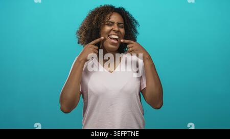 Frau mit lockigen Haaren in rosa T-Shirt zeigt Finger zu Mund und zeigt Zähne und ein breites Lächeln im türkisfarbenen Studio; Freude. Stockfoto