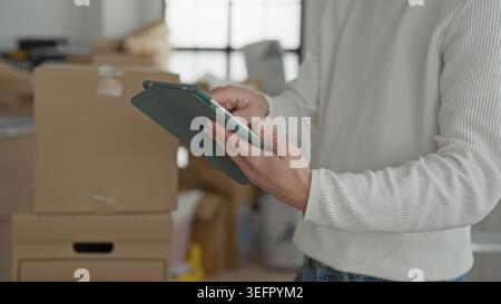 Mann, der Tablets hält und gestapelte Umzugskartons und Pakete im Gebäude überprüft; fokussierte Bestandsverwaltung. Stockfoto