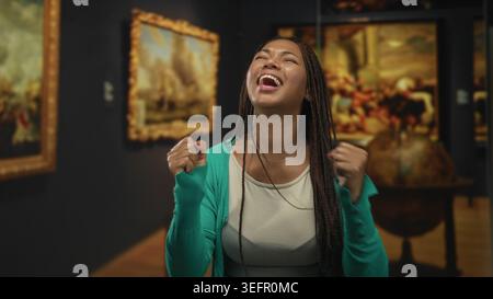 Frau lacht mit geballten Fäusten in einem Kunstgalerie-Gebäude, geflochtene Haare und grüne Strickjacke sichtbar; Freude feiern Wunder. Stockfoto