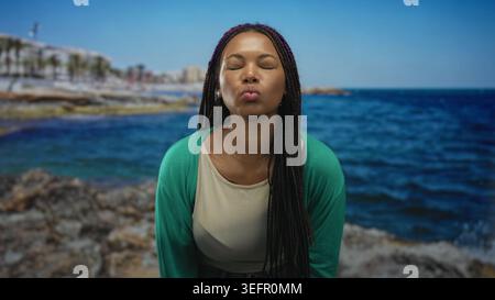 Frau spitzt die Lippen mit geschlossenen Augen auf Felsen am Meer und zeigt die Zeigefinger nach oben; spielerisches Selbstvertrauen. Stockfoto