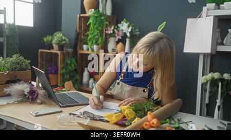 Blonde Floristin mit Schürze neben Laptop und Blumenstrauß in einem Floristikstudio; fokussierte Planung. Stockfoto