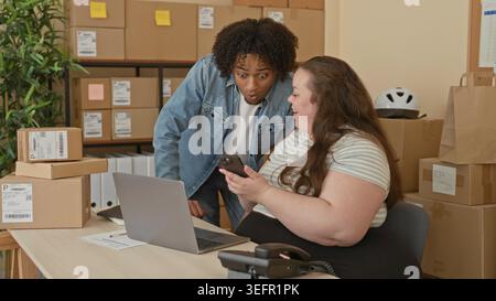 Mann und Frau tauschen einen hohen fünf aus, während sie ein Smartphone neben einem Laptop halten und Versandkartons in einem Gebäude stapeln Stockfoto