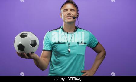 Der Schiedsrichter hält den Fußball in gestreckter Hand, mit Pfeife und Headset vor der violetten Studiowand; Selbstvertrauen. Stockfoto