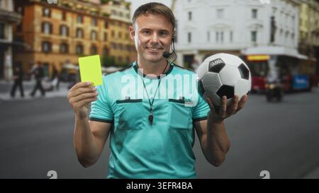 Schiedsrichter hält gelbe Karte und Fußball auf der Straße, mit Pfeife und Headset; Fairplay-Selbstvertrauen. Stockfoto