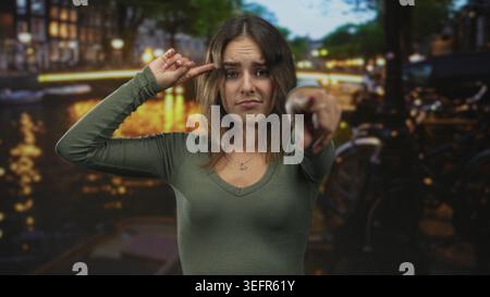 Die junge hispanische Frau zeigt mit dem Finger auf die Kamera und klopft in der Abenddämmerung an den Tempel einer Grachtenstraße in amsterdam. Stockfoto