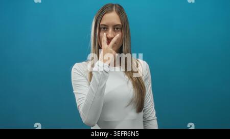 Junge hispanische Frau, die Hand zeigt, die von der Hüfte nach oben in einem Studio mit blaublauem Hintergrund und neutralem Ausdruck auf die Kamera zeigt; Entschlossenheit. Stockfoto