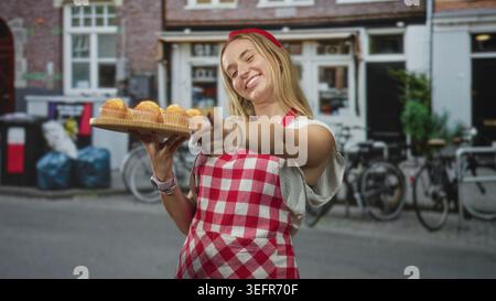 Die Chefin hält ein hölzernes Tablett mit Muffins und zeigt mit dem Finger, während sie auf der Straße eine rote Gingham-Schürze trägt; gastfreundliche Gastfreundschaft. Stockfoto