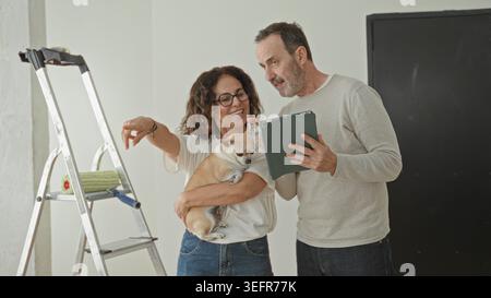 Der Mann hält ein Tablet, während die Frau einen kleinen Hund in die Wiege legt und mit der rechten Hand in Richtung Leiter und Malerwalze in einem Gebäude zeigt; Zufriedenheit bei der Renovierung des Hauses. Stockfoto