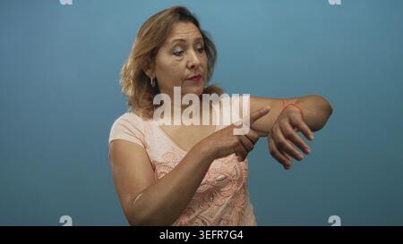 hispanische Frau mittleren Alters zeigt Finger an Handgelenk gegen blaues Studio-Setting; Ungeduld Zeitbewusstsein. Stockfoto