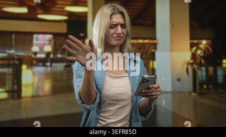 Frau runzelte die Stirn, während sie ihr Smartphone hält und Nachrichten in der Lobby des Gebäudes mit Lampen und Sitzgelegenheiten liest; frustrierende Kommunikation. Stockfoto
