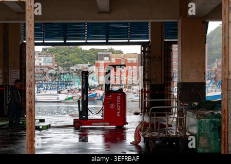 Yilan, Taiwan - 31. März 2025: Roter Gabelstapler im Nanfangao Fischmarkt mit farbenfrohen Fischerbooten und Hafen, sichtbar durch den offenen Eingang. Stockfoto