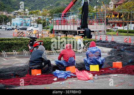Yilan, Taiwan - 31. März 2025: Drei Wanderarbeiter reparieren Fischernetze auf dem Gehweg in Nanfangao, mit einem Kranwagen und einem Tempel im Hintergrund. Stockfoto