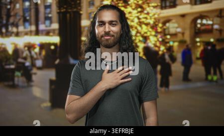 Junger Mann mit rechter Hand auf der Brust auf der Straße vor dem beleuchteten Weihnachtsbaum und verschwommenen Einkäufern, kurze Halskette sichtbar, graues T-Shirt trägt; Dankbarkeit. Stockfoto