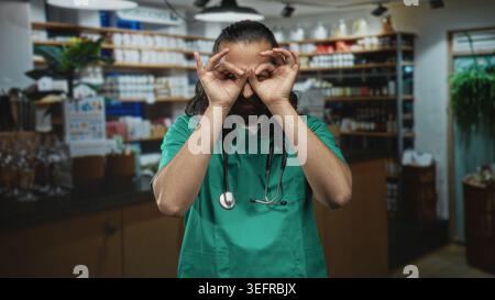 Mann mit grünen Peelings und Stethoskop lächelnd mit Händen als Fernglas beim Bau eines Apothekengangs; freundliches Pflegevertrauen. Stockfoto