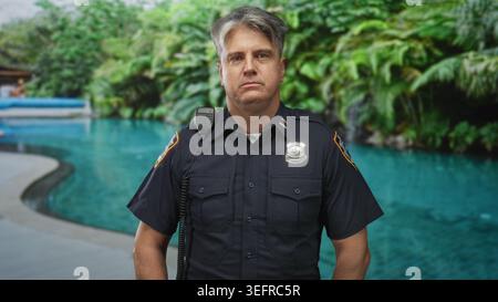 Polizist in Uniform am Poolrand mit Funkmikrofon an der Schulter und Abzeichen sichtbar, neutraler Ausdruck, Gebäude; ernste Pflicht. Stockfoto