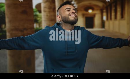 Mann mit blauem Kapuzenpullover mit ausgestreckten Armen im Gang des Altstadtgebäudes, lächelnd mit geschlossenen Augen; Freude Freiheit. Stockfoto
