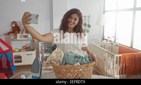Frau, die Wäschekorb und Bügeleisen auf dem Bügelbrett hält, während sie lächelt und ein Selfie im Haus macht; Mütterlichkeit Routine Freude. Stockfoto