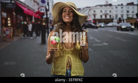 Frau mit zwei Eiskegeln in der Hand auf einer belebten Straße, die von Geschäften und Autos gesäumt ist, mit Strohhut und gelbem Oberteil; verspielter Sommer. Stockfoto