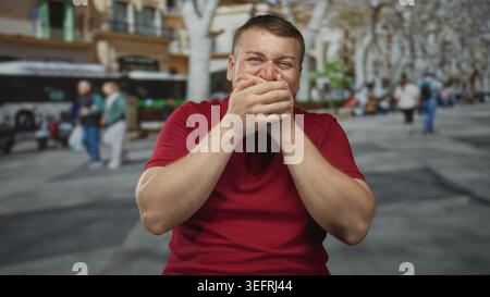 Mann in rotem Hemd, der den Mund mit beiden Händen auf einer überfüllten Stadtstraße in der Nähe eines Busses bedeckt und Fußgänger läuft; Lachen. Stockfoto
