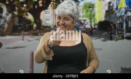 Frau zeigt auf der Straße den Finger auf die Kamera, lächelt mit sichtbaren Fingern und Armbändern, trägt Strickjacke und schwarzes Tanktop; verspielte Ermutigung. Stockfoto