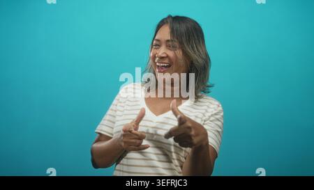 Frau zeigt beide Zeigefinger in Fingerpistolen, während sie im Studio mit türkisfarbener Kulisse lacht und lächelt; fröhliche Verspieltheit. Stockfoto