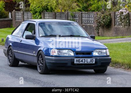 Ein klassischer, blauer Ford Sierra 1990 dreitüriger Fließheck mit speziell angefertigten Leichtmetallrädern fährt auf dem Weg zur Renntag Car Show 2026 auf einer Wohnstraße in Großbritannien. Stockfoto