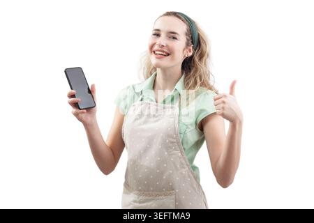 Eine lächelnde Hausfrau, die eine Kochschürze trägt, hält ein Smartphone und zeigt die Daumen hoch. Isoliert auf weißem Hintergrund, für Food-Apps oder zum Kochen Stockfoto