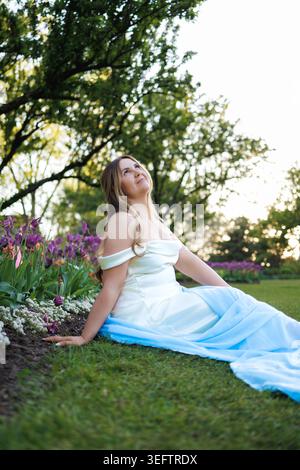 Eine Frau genießt Zeit in einem Garten voller Blumen, in einem hellblauen Kleid. Sie sitzt auf dem Gras und blickt in den Himmel, während die Sonne untergeht und baden Stockfoto