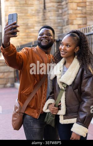 Afroamerikanisches Paar nimmt Smartphone-Selfie, lacht auf der Straße in Mänteln, Umhängetaschen Stockfoto