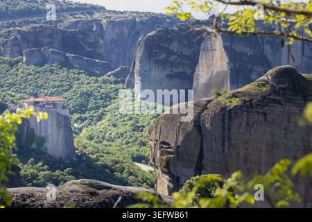 Fantastisches meteora-Kloster, das auf massiven Klippen mit einem weitläufigen Tal thront, ein dramatischer Blick auf das unesco-Weltkulturerbe durch natürliche Blätter im Vordergrund Stockfoto