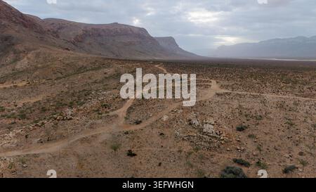 Gewundene Feldstraße durch felsige Wüstenebene mit weit entfernten Bergen unter bewölktem Himmel, weite Luftlandschaften, abgelegenes Reisekonzept in der Wildnis. Stockfoto