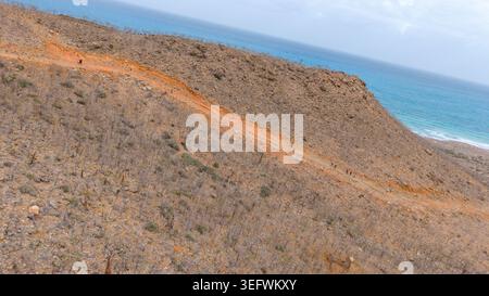 Aus der Vogelperspektive auf den trockenen Küsten-Hügel mit orangefarbenem Feldweg entlang des Berges und blauem Ozean im Hintergrund unter klarem Himmel. Stockfoto