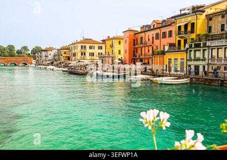 Peschiera del Garda, Italien - 30. April 2026: Lebhafte Gebäude säumen den Kanal in Peschiera del Garda, Italien, mit angedockten Booten und Blumen in Töpfen Stockfoto