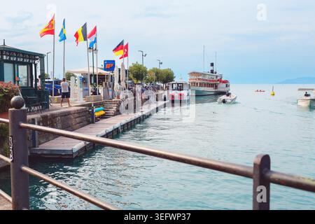 Peschiera Del Garda, Italien - 30. April 2026: Blick auf das Ufer von Peschiera del Garda mit Booten am Dock, farbenfrohen Flaggen und Besuchern Stockfoto