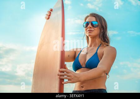 Selbstbewusste junge Frau in Bikini und Sonnenbrille, die ein Surfbrett hält und draußen unter einem klaren blauen Himmel steht. Die Ansicht mit niedrigem Winkel betont die Stärke, die frei ist Stockfoto