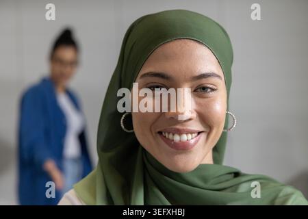 Zwei weibliche Kollegen lächelten im Büro in die Kamera, eine Frau mit grünem Kopftuch und Ohrringen Stockfoto