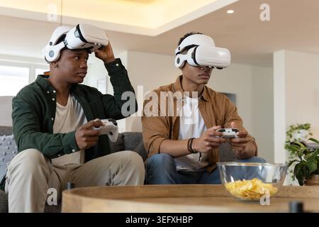 Verschiedene männliche Freunde sitzen im Wohnzimmer auf dem Sofa und tragen weiße VR-Headsets mit Controllern Stockfoto