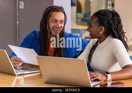 Verschiedene Kollegen sitzen am Holztisch mit blauer Jacke und weißem Oberteil und halten Papier an Laptops Stockfoto