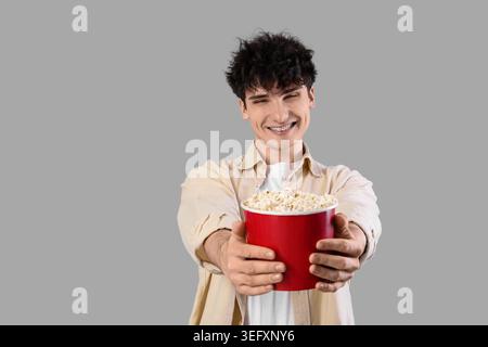 Junger Mann, der Popcorn-Eimer auf grauem Hintergrund hält Stockfoto