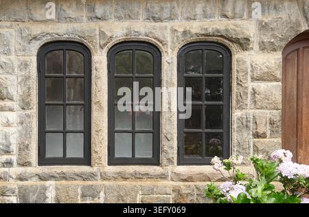 Schwarze bogenförmige Glasfenster an rustikaler grauer Steinwand, mehrschichtige Rahmen mit reflektierendem Glas und blühenden Blumen, Seitenansicht des alten europäischen zeitalters Stockfoto