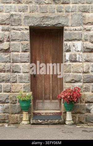 Vintage braune Holztür auf grauer Steinblockwand, symmetrischer Eingang des Häuschens mit Blumentöpfen, Seitenansicht des traditionellen Wohnhauses Stockfoto