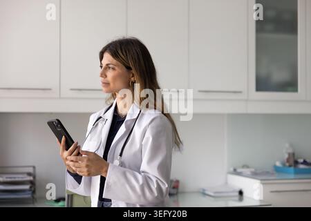 Nachdenkliche Ärztin im weißen Mantel mit Smartphone am Arbeitsplatz Stockfoto
