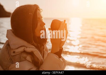 Frau Apfel Sonnenuntergang ruhige junge Frau, die einen warmen Hoodie trägt, hält einen frischen Apfel am Strand während der goldenen Stunde Stockfoto