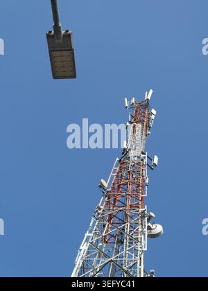 Flacher Blick auf ein rot-weißes Telekommunikationsnetz mit mehreren Antennen und Mikrowellenschalen vor klarem blauem Himmel, mit einer Straße Stockfoto