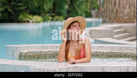 Junge Frau in Strohhut und farbenfrohen Badeanzügen, die sich in einem luxuriösen Resort-Swimmingpool mit üppigem tropischem Grün entspannen. Ein Konzept für Luxusurlaub, Stockfoto