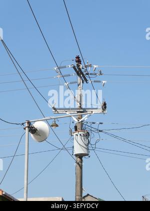 Versorgungsmast aus Holz mit zylindrischem Transformator, Keramikisolatoren und mehreren Überlandstromkabeln neben einer runden Straßenlaterne unter einem klaren B Stockfoto